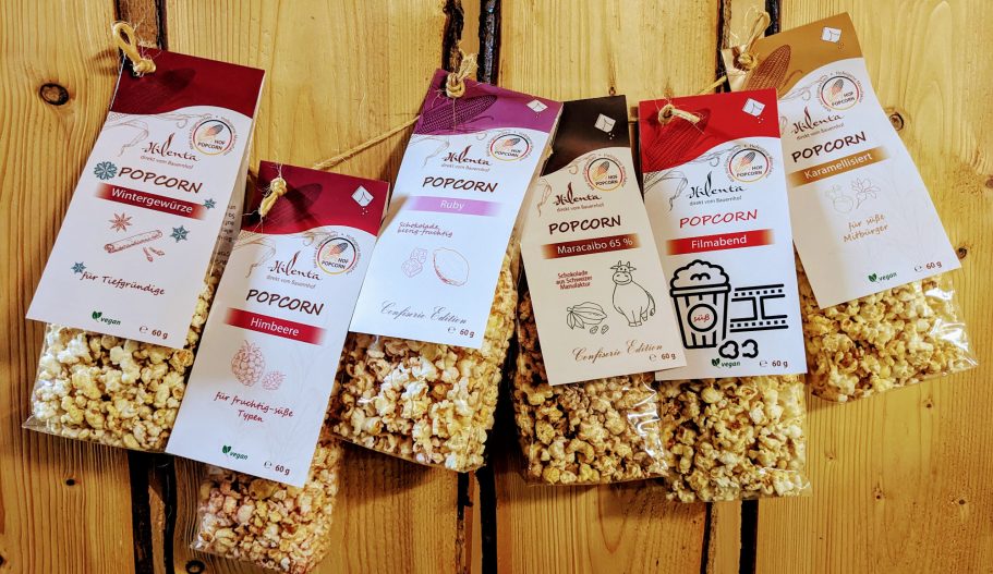 Sechs Tüten gepuffter Snacks mit bunten Etiketten an einer Holzwand aufgehängt.